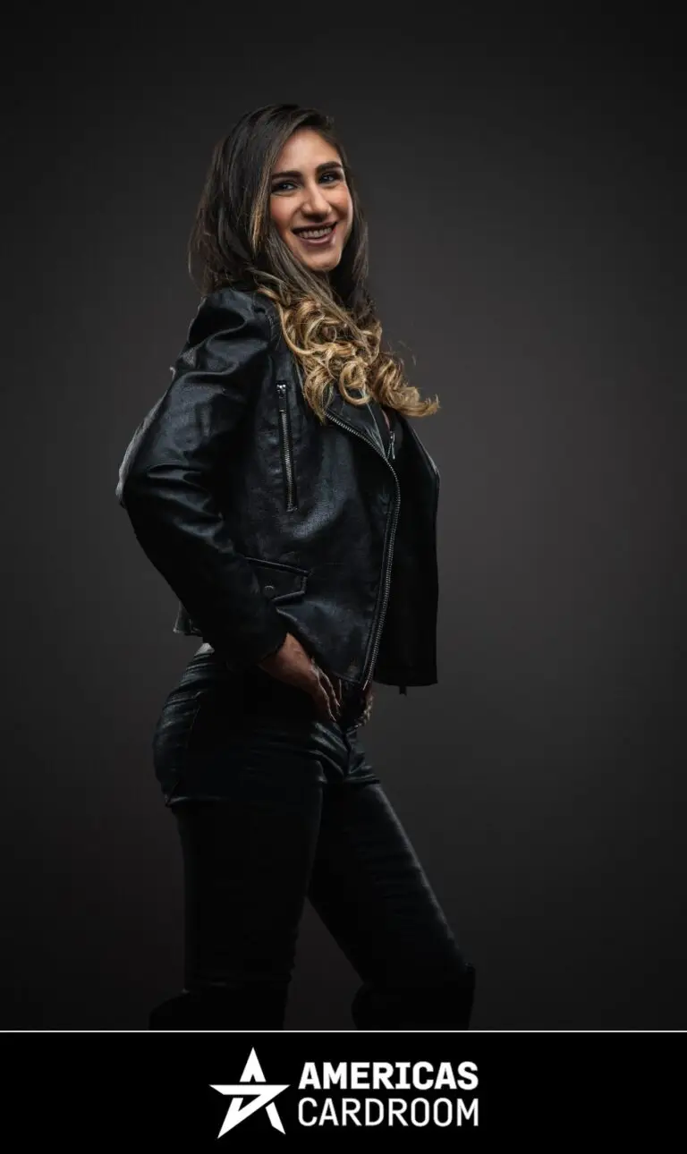 A three-quarter profile shot of Ana Marquez, an ACR poker pro, smiling and looking back towards the camera. She has long, dark hair with blonde highlights and is wearing a black leather motorcycle jacket and black shiny pants, with her hands on her hips, against a dark background.