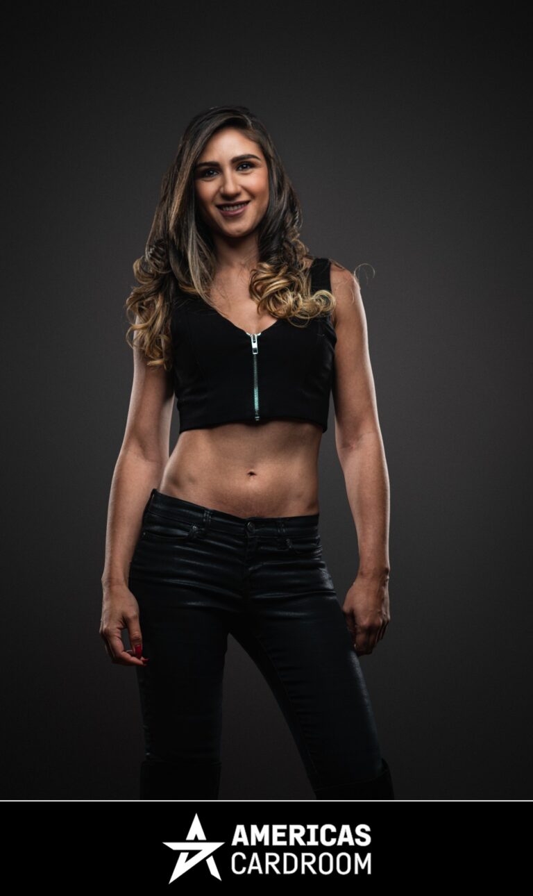 A medium shot of Ana Marquez, an ACR poker pro, standing with a slight smile. She has long, dark hair with lighter highlights and is wearing a black sleeveless crop top with a visible green zipper, and black shiny pants, against a dark background. Her fingernails are painted red.