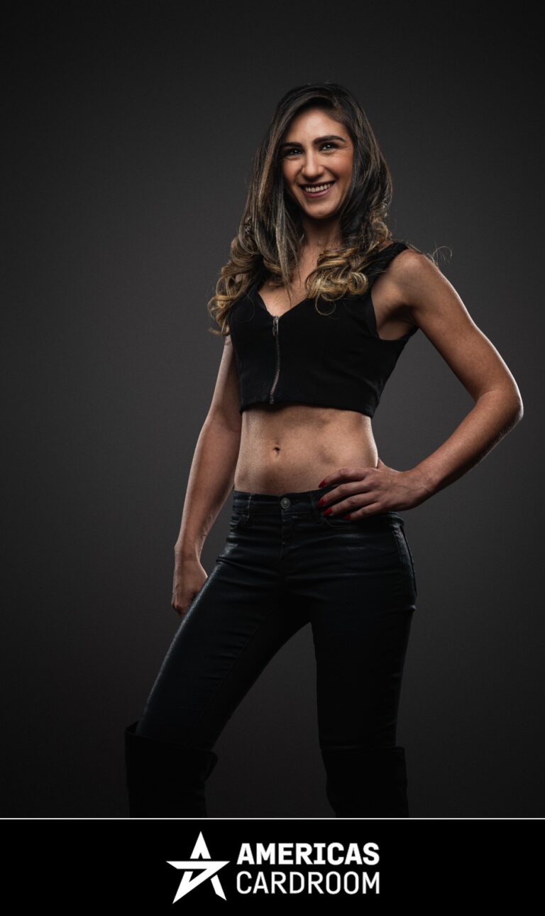 A medium shot of Ana Marquez, an ACR poker pro, standing with her left hand on her hip and a wide smile. She has long, dark hair with lighter highlights and is wearing a black sleeveless crop top with a zipper down the front, and black shiny pants, against a dark background. Her fingernails are painted red.