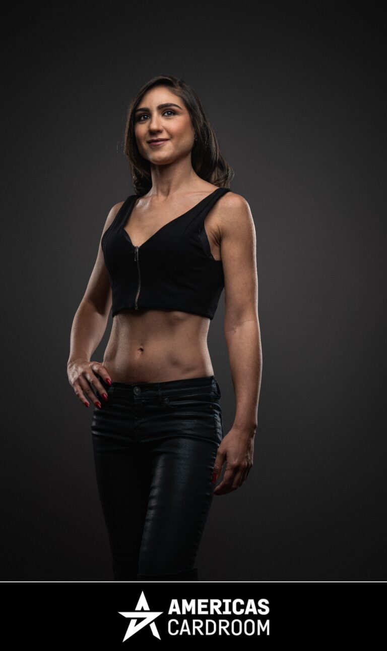 A medium shot of Ana Marquez, an ACR poker pro, standing with her left hand on her hip and a slight smile. She has long, dark hair and is wearing a black sleeveless crop top with a zipper down the front, and black shiny pants, against a dark background. Her fingernails are painted red.