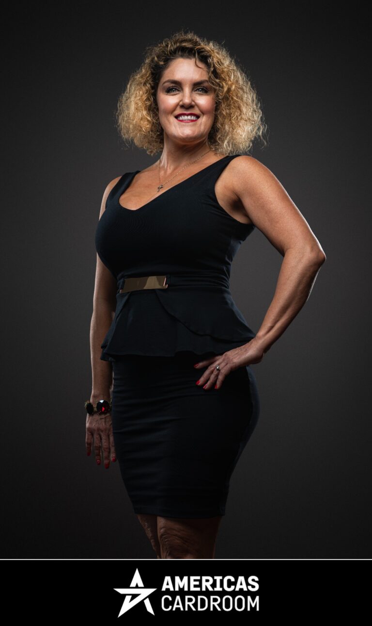 A medium shot of Casey Nezhoda, an ACR poker pro, with curly blonde hair, standing with her right hand on her hip and a wide smile. She is wearing a black sleeveless dress with a V-neck and a peplum waist, cinched with a gold belt, against a dark background. She has red fingernails and is wearing a ring on her left hand and bracelets on her right wrist.