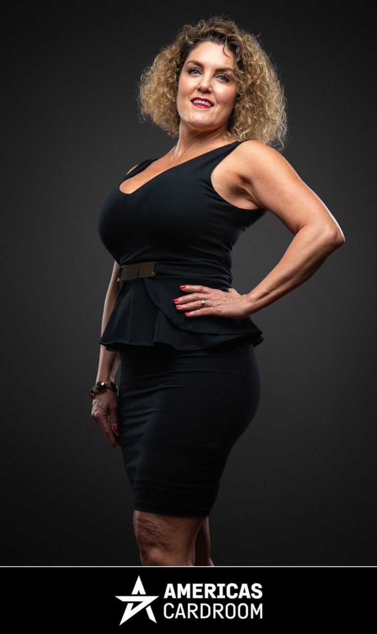 A medium shot of Casey Nezhoda, an ACR poker pro, with curly blonde hair, standing with her right hand on her hip and a wide smile. She is wearing a black sleeveless dress with a V-neck and a peplum waist, cinched with a gold belt, against a dark background. She has red fingernails and is wearing a ring on her left hand and bracelets on her right wrist.
