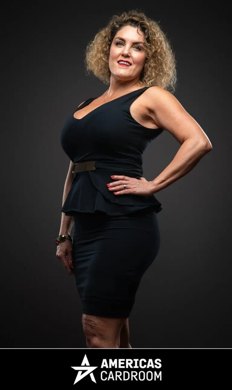 A medium shot of Casey Nezhoda, an ACR poker pro, with curly blonde hair, standing with her right hand on her hip and a wide smile. She is wearing a black sleeveless dress with a V-neck and a peplum waist, cinched with a gold belt, against a dark background. She has red fingernails and is wearing a ring on her left hand and bracelets on her right wrist.