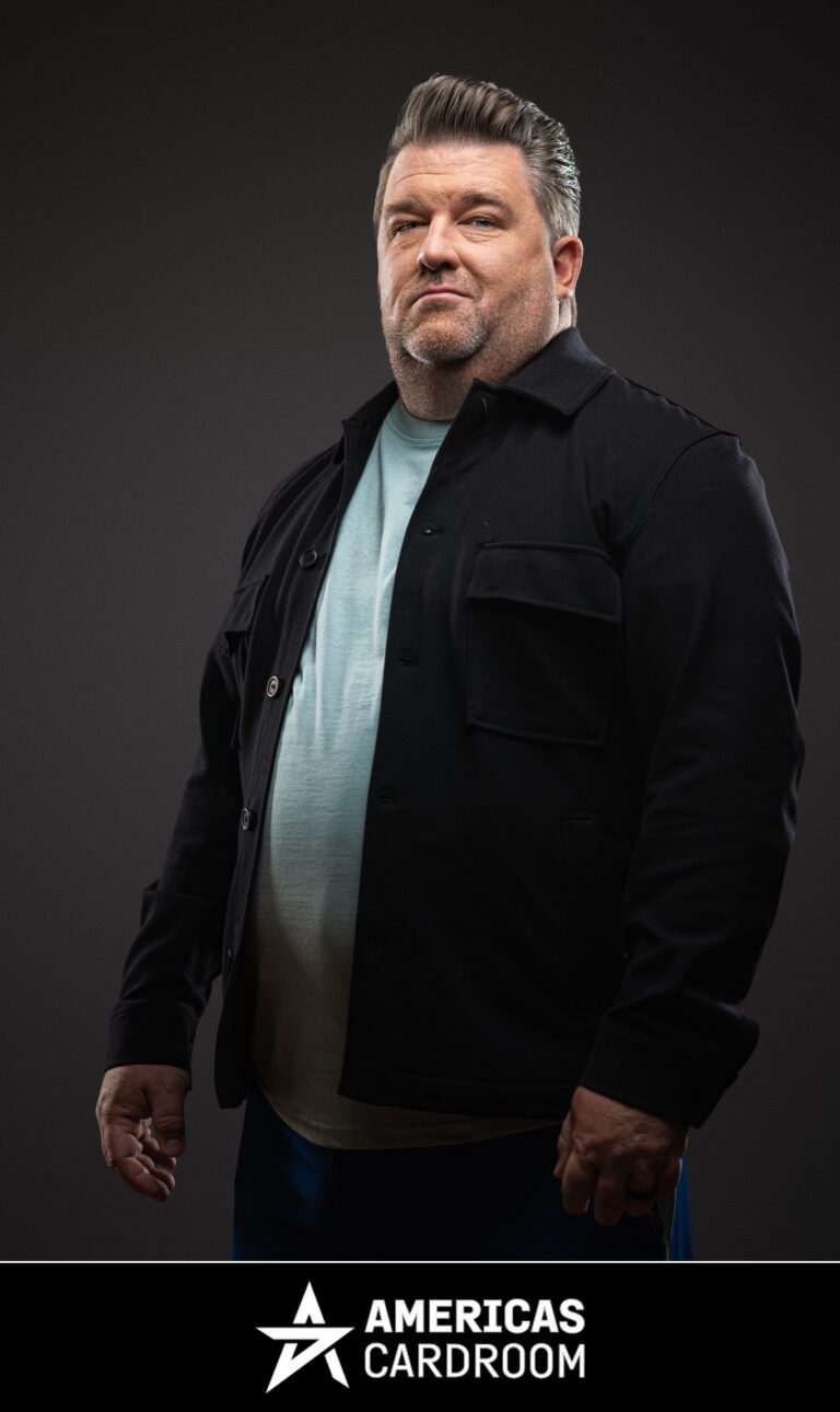 A medium shot of Chris Moneymaker, an ACR poker pro, with a beard, looking to his left with a neutral expression. He is wearing a black button-up jacket open over a light blue t-shirt, and blue pants, against a dark background.