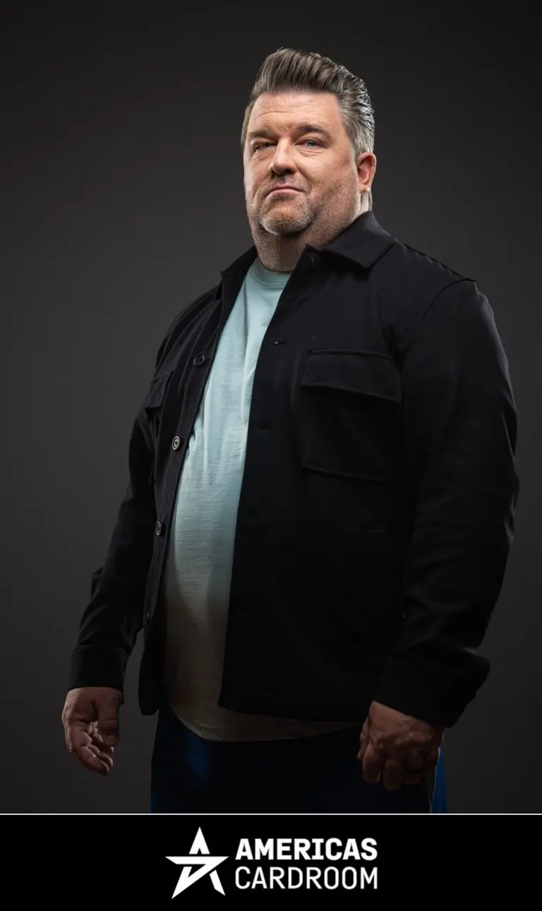 A medium shot of Chris Moneymaker, an ACR poker pro, with a beard, looking to his left with a neutral expression. He is wearing a black button-up jacket open over a light blue t-shirt, and blue pants, against a dark background.