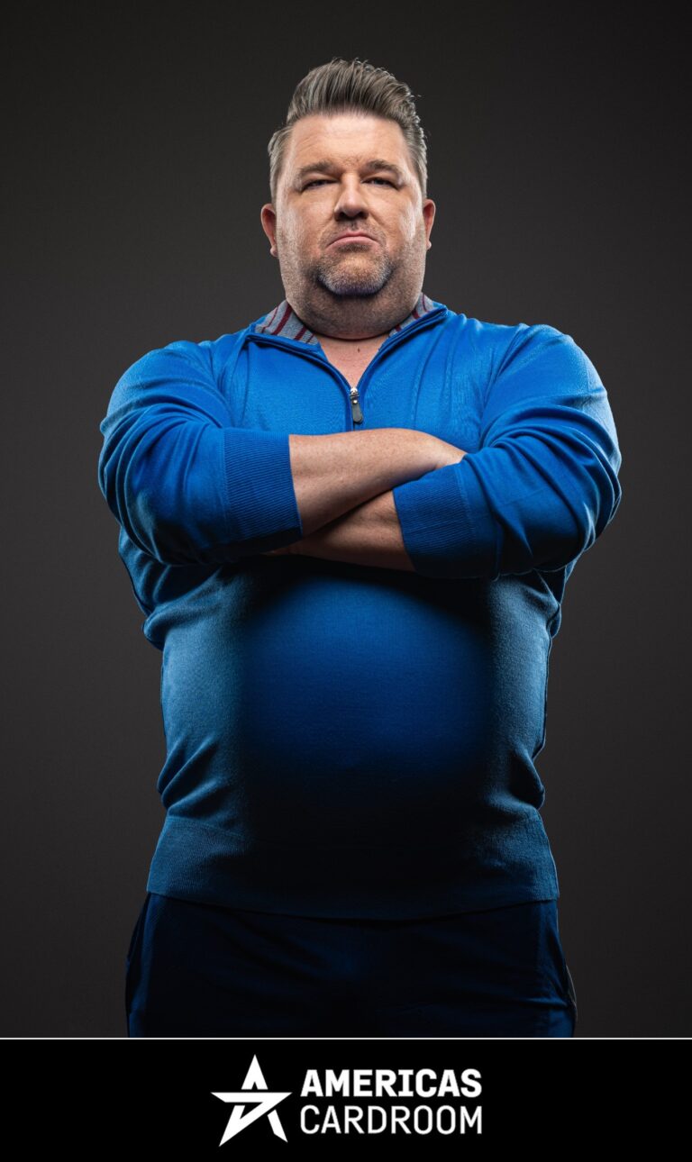 A medium shot of Chris Moneymaker, an ACR poker pro, with a beard, looking directly at the camera with a serious expression and his arms crossed. He is wearing a blue zip-up sweater and dark pants, against a dark background.