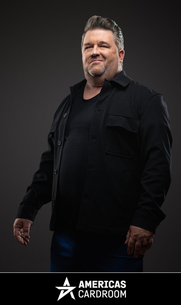 A medium shot of Chris Moneymaker, an ACR poker pro, with a beard, looking to his right with a slight smile. He is wearing a black button-up jacket over a black t-shirt and blue pants, against a dark background.