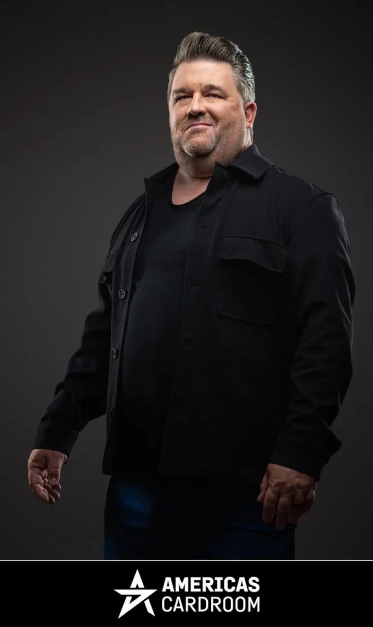A medium shot of Chris Moneymaker, an ACR poker pro, with a beard, looking to his right with a slight smile. He is wearing a black button-up jacket over a black t-shirt and blue pants, against a dark background.