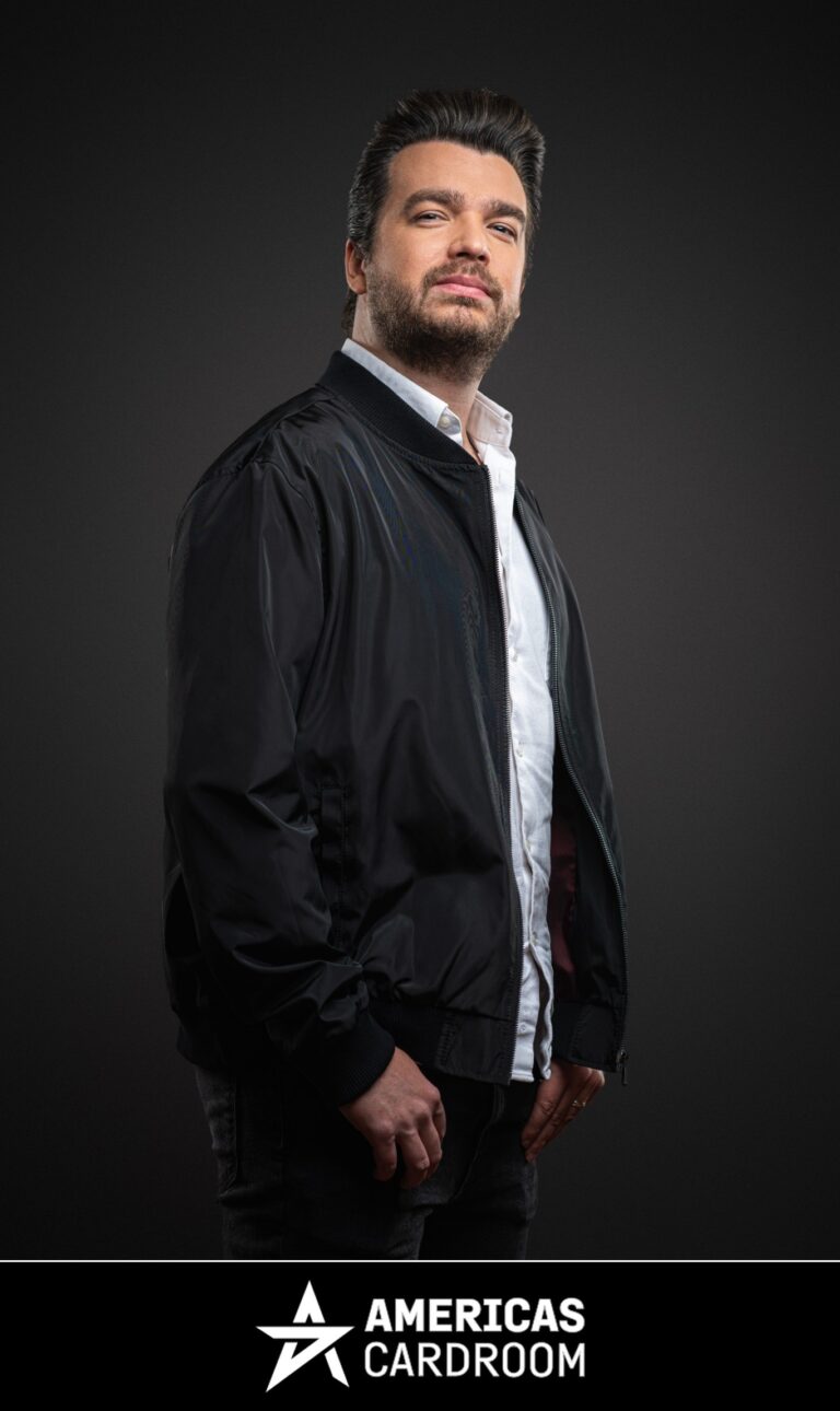 A medium shot of Chris Moorman, an ACR poker pro, with dark hair and a beard, looking directly at the camera with a slight smile. He is wearing a black bomber jacket open over a white collared shirt and dark pants, against a dark background.