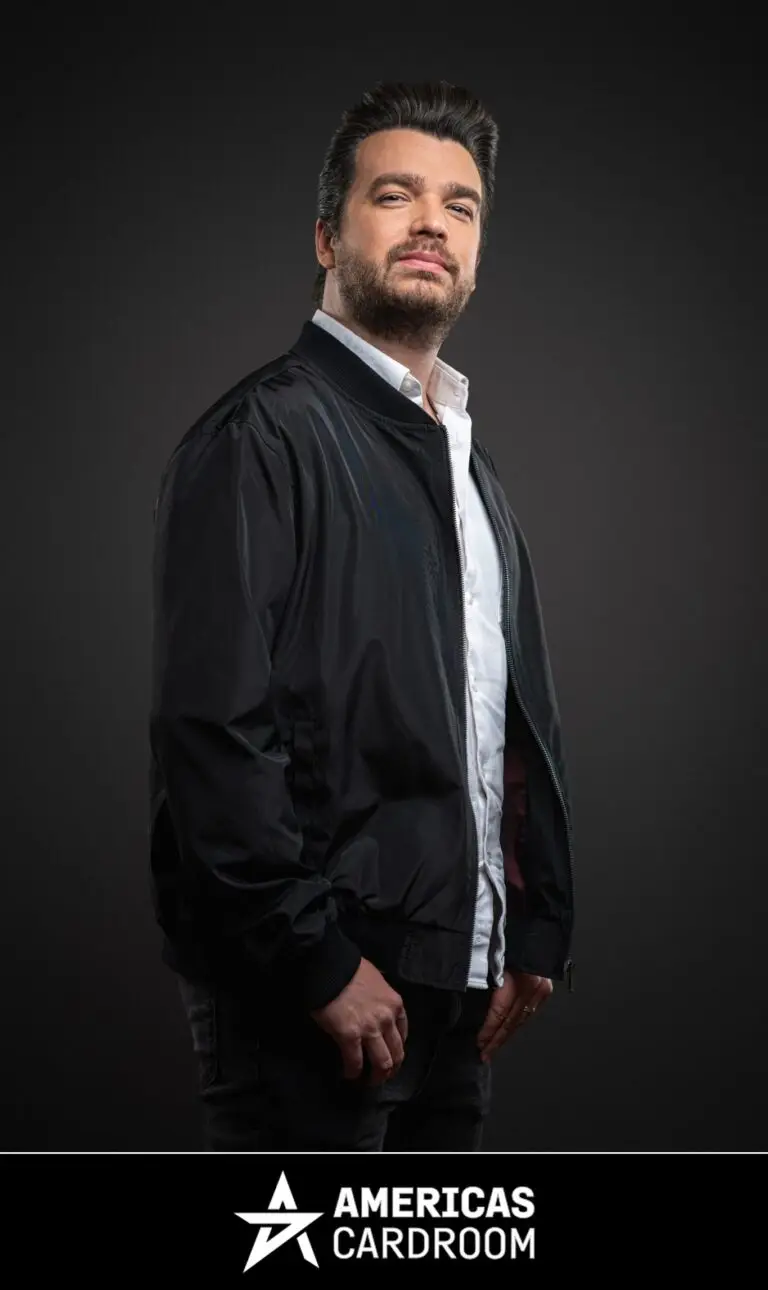 A medium shot of Chris Moorman, an ACR poker pro, with dark hair and a beard, looking directly at the camera with a slight smile. He is wearing a black bomber jacket open over a white collared shirt and dark pants, against a dark background.