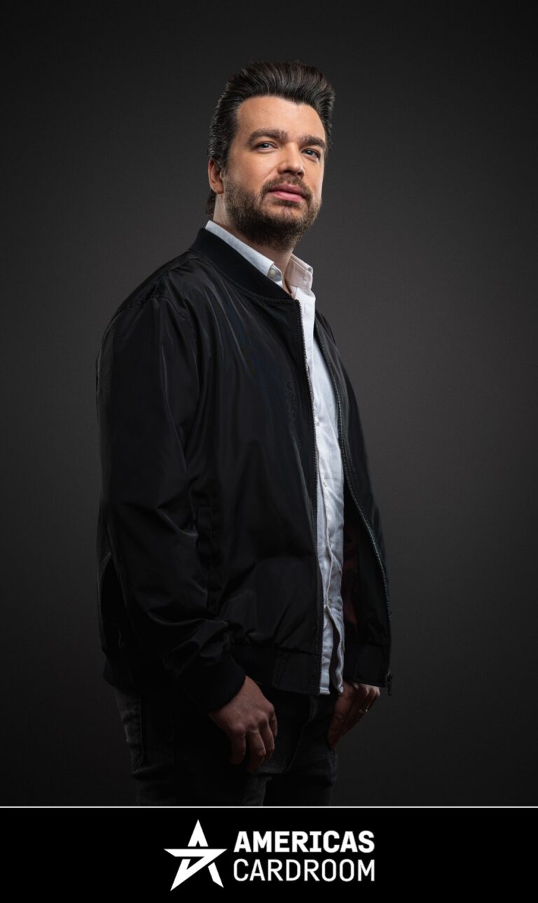 A medium shot of Chris Moorman, an ACR poker pro, with dark hair and a beard, looking directly at the camera with a serious expression. He is wearing a black bomber jacket open over a white collared shirt and dark pants, against a dark background.