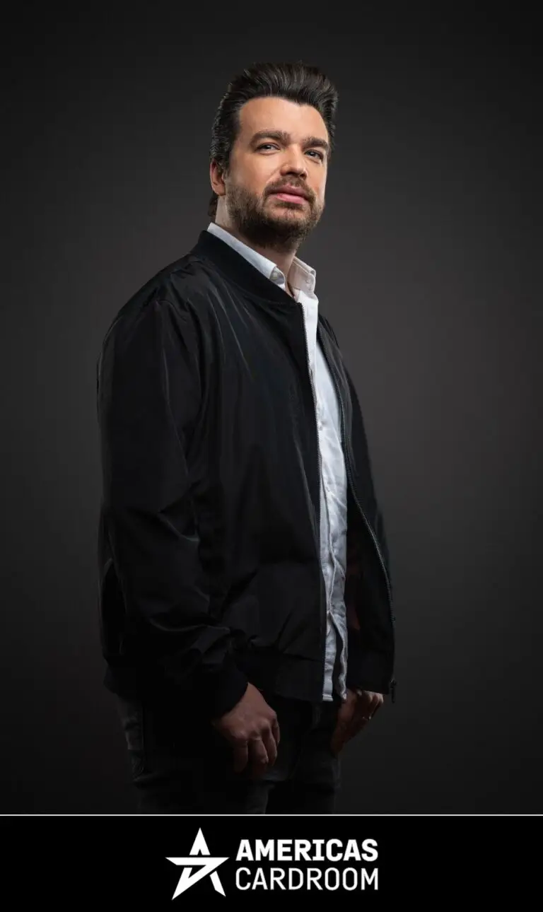A medium shot of Chris Moorman, an ACR poker pro, with dark hair and a beard, looking directly at the camera with a serious expression. He is wearing a black bomber jacket open over a white collared shirt and dark pants, against a dark background.