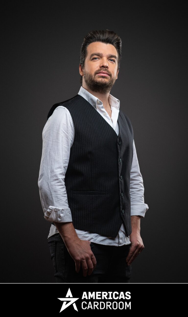 A medium shot of Chris Moorman, an ACR poker pro, with dark hair and a beard, looking directly at the camera with a serious expression. He is wearing a dark pinstriped vest over a white long-sleeved shirt with rolled-up sleeves, and dark pants, against a dark background.