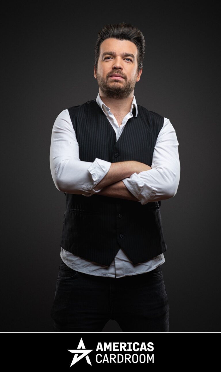 A medium shot of Chris Moorman, an ACR poker pro, with dark hair and a beard, looking directly at the camera with a serious expression and his arms crossed. He is wearing a dark pinstriped vest over a white long-sleeved shirt with rolled-up sleeves, and dark pants, against a dark background.