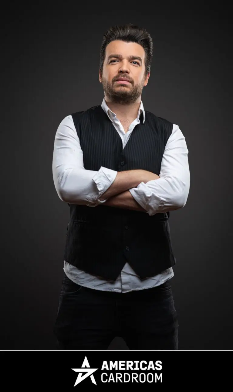 A medium shot of Chris Moorman, an ACR poker pro, with dark hair and a beard, looking directly at the camera with a serious expression and his arms crossed. He is wearing a dark pinstriped vest over a white long-sleeved shirt with rolled-up sleeves, and dark pants, against a dark background.