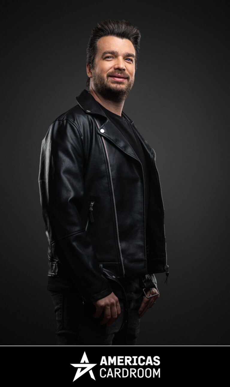 A medium shot of Chris Moorman, an ACR poker pro, with dark hair and a beard, smiling and looking to his left. He is wearing a black leather motorcycle jacket over a black t-shirt and dark pants, against a dark background.
