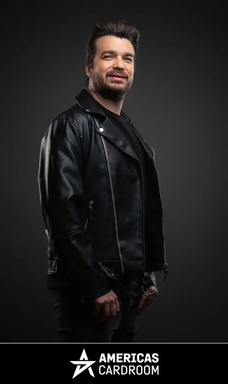 A medium shot of Chris Moorman, an ACR poker pro, with dark hair and a beard, smiling and looking to his left. He is wearing a black leather motorcycle jacket over a black t-shirt and dark pants, against a dark background.