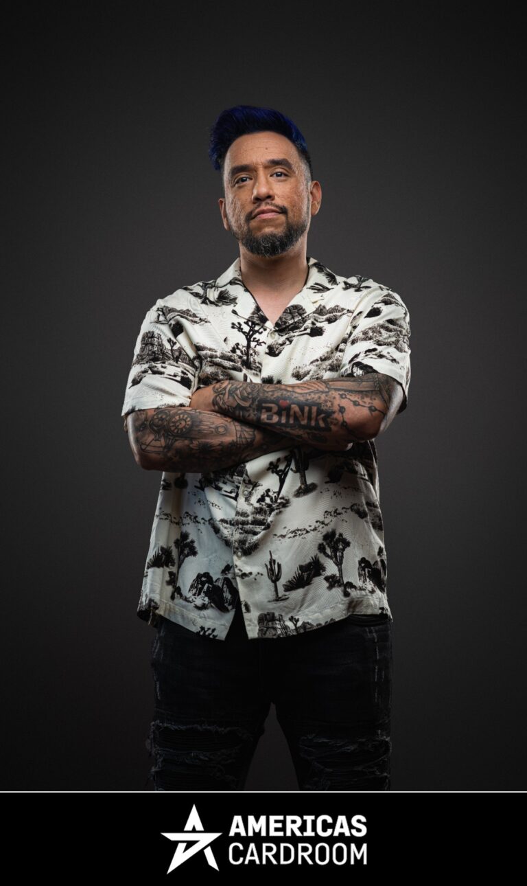 A medium shot of Drew Gonzalez, an ACR poker pro, with blue-tipped hair and a beard, standing with his arms crossed and a serious expression. He is wearing a white short-sleeved button-up shirt with a black desert-themed pattern, and dark jeans, with tattoos visible on his arms, against a dark background.