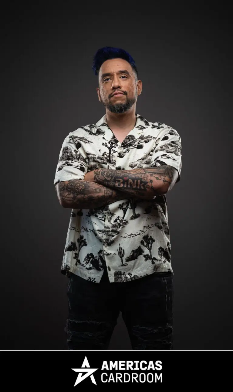 A medium shot of Drew Gonzalez, an ACR poker pro, with blue-tipped hair and a beard, standing with his arms crossed and a serious expression. He is wearing a white short-sleeved button-up shirt with a black desert-themed pattern, and dark jeans, with tattoos visible on his arms, against a dark background.
