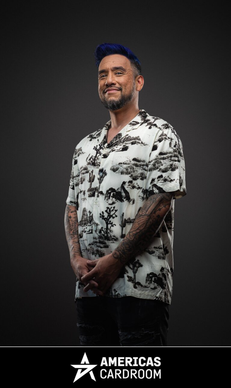 A medium shot of Drew Gonzalez, an ACR poker pro, with blue-tipped hair and a beard, smiling and looking to his left. He is wearing a white short-sleeved button-up shirt with a black desert-themed pattern, and dark jeans, with his hands clasped in front of him, and tattoos visible on his arms, against a dark background.