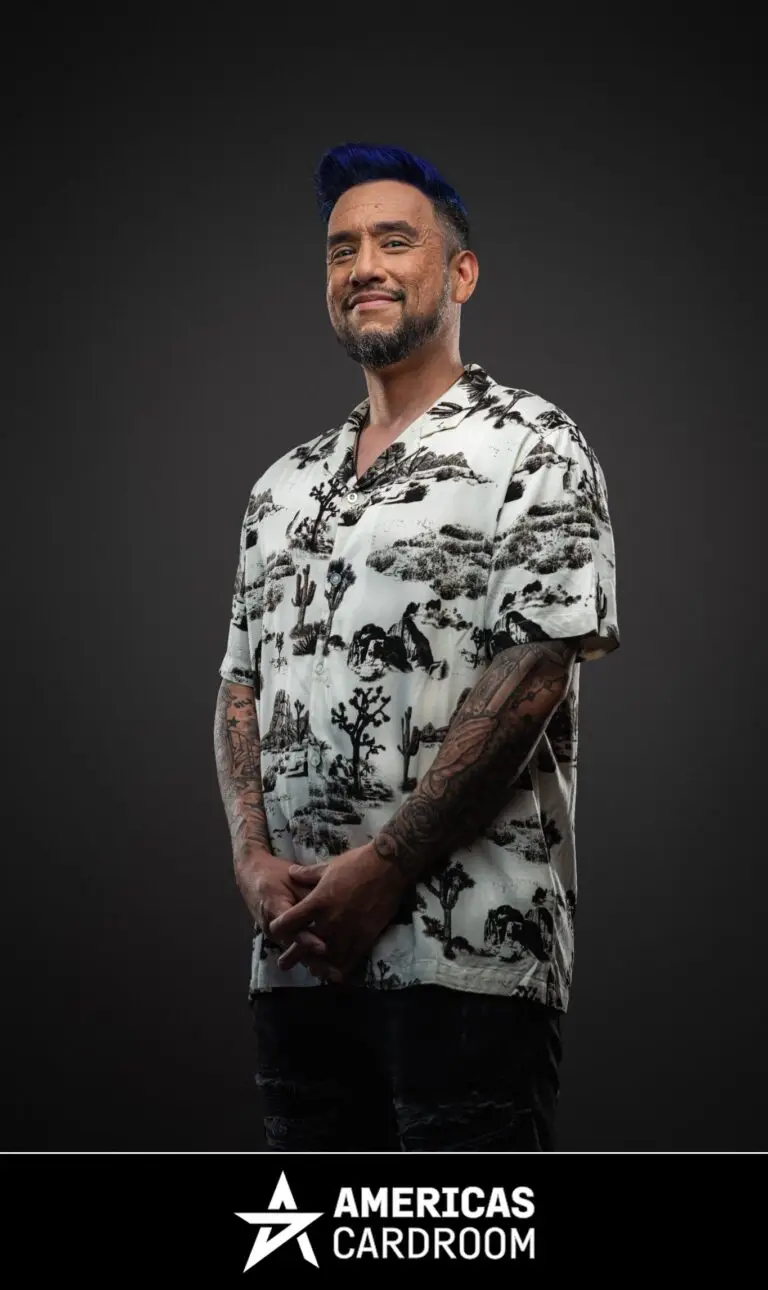 A medium shot of Drew Gonzalez, an ACR poker pro, with blue-tipped hair and a beard, smiling and looking to his left. He is wearing a white short-sleeved button-up shirt with a black desert-themed pattern, and dark jeans, with his hands clasped in front of him, and tattoos visible on his arms, against a dark background.