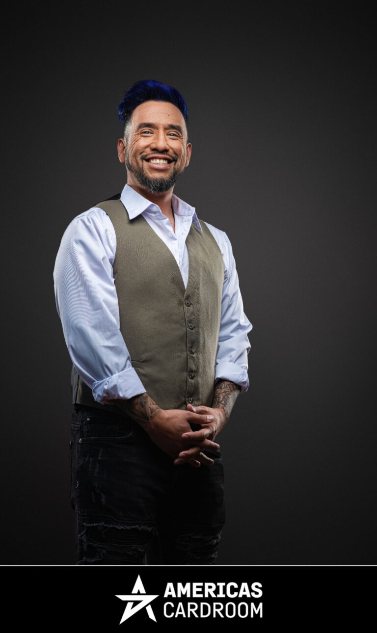 A medium shot of Drew Gonzalez, an ACR poker pro, with blue-tipped hair and a beard, smiling widely and looking to his left. He is wearing a grey vest over a light blue collared shirt with rolled-up sleeves, and dark jeans, with his hands clasped in front of him, and tattoos visible on his arms, against a dark background.