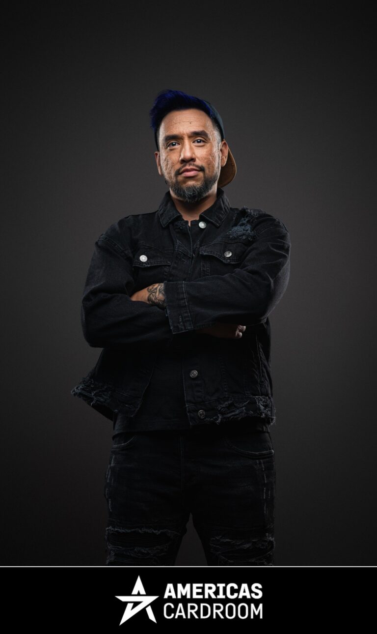 A medium shot of Drew Gonzalez, an ACR poker pro, with a beard and a blue baseball cap worn backward. He is standing with his arms crossed and a serious expression, wearing a black denim jacket and black ripped jeans, with tattoos visible on his left arm, against a dark background.