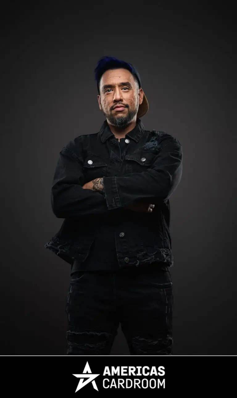 A medium shot of Drew Gonzalez, an ACR poker pro, with a beard and a blue baseball cap worn backward. He is standing with his arms crossed and a serious expression, wearing a black denim jacket and black ripped jeans, with tattoos visible on his left arm, against a dark background.