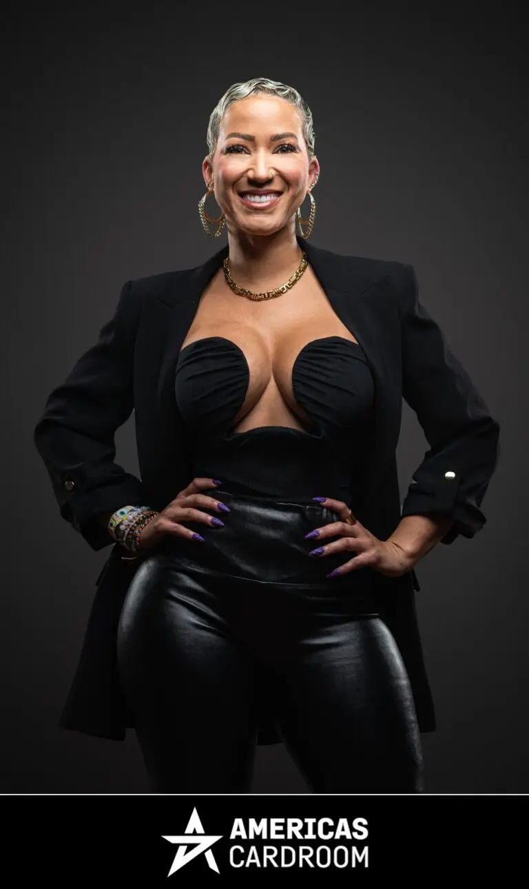 A medium shot of Ebony Kenney, an ACR poker pro, with short, styled hair, standing with her hands on her hips and a wide smile. She is wearing a black blazer over a black strapless top with a plunging neckline, black shiny pants, large hoop earrings, and a gold chain necklace, against a dark background. She also has colorful bracelets on her left wrist. Her fingernails are painted purple.