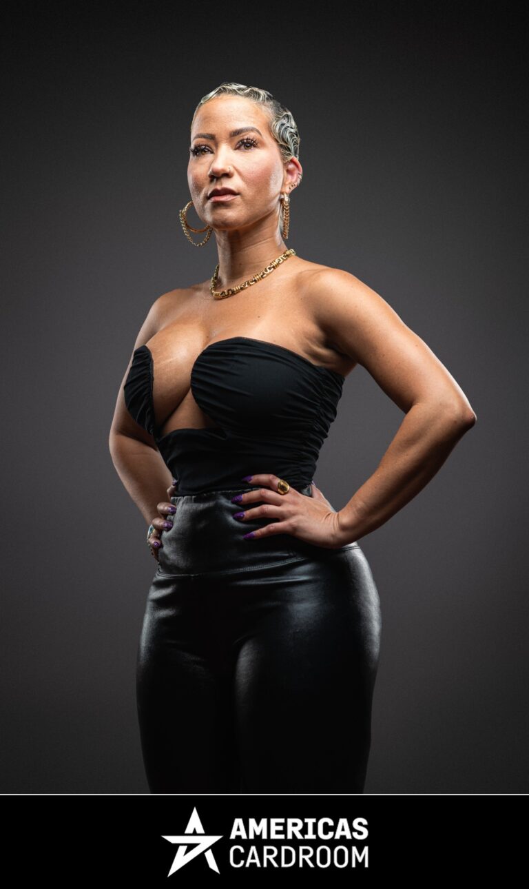 A medium shot of Ebony Kenney, an ACR poker pro, with short, styled hair, standing with her hands on her hips and a confident expression. She is wearing a black strapless top with a plunging neckline, black shiny pants, large hoop earrings, and a gold chain necklace, against a dark background. Her fingernails are painted purple.