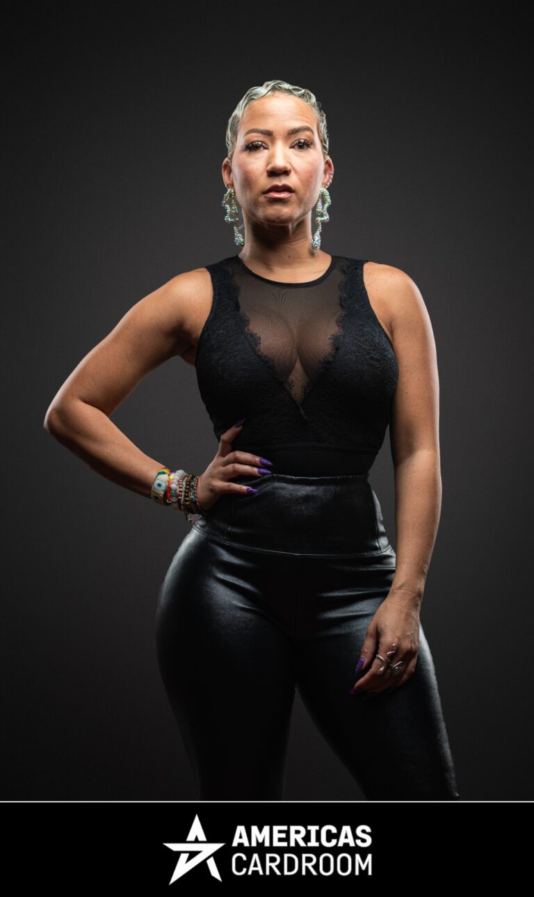 A medium shot of Ebony Kenney, an ACR poker pro, with short, styled hair, standing with her right hand on her hip and a serious expression. She is wearing a black sleeveless top with a sheer and lace chest panel, black shiny pants, large drop earrings, and colorful bracelets on her left wrist, against a dark background. Her fingernails are painted purple.