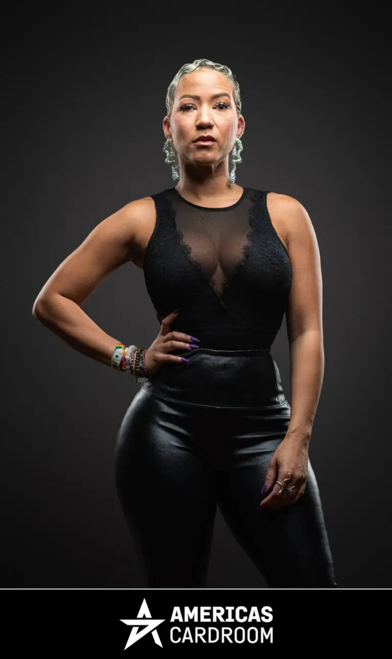 A medium shot of Ebony Kenney, an ACR poker pro, with short, styled hair, standing with her right hand on her hip and a serious expression. She is wearing a black sleeveless top with a sheer and lace chest panel, black shiny pants, large drop earrings, and colorful bracelets on her left wrist, against a dark background. Her fingernails are painted purple.