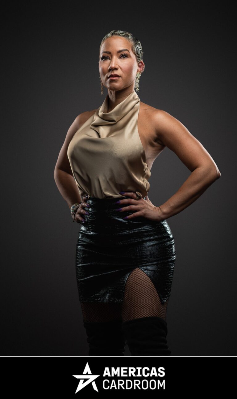 A medium shot of Ebony Kenney, an ACR poker pro, with short, styled hair, standing with her hands on her hips and a confident expression. She is wearing a gold sleeveless cowl-neck top, a black shiny skirt with a high slit revealing fishnet stockings, and knee-high black boots, against a dark background. She is also wearing large earrings and a bracelet.