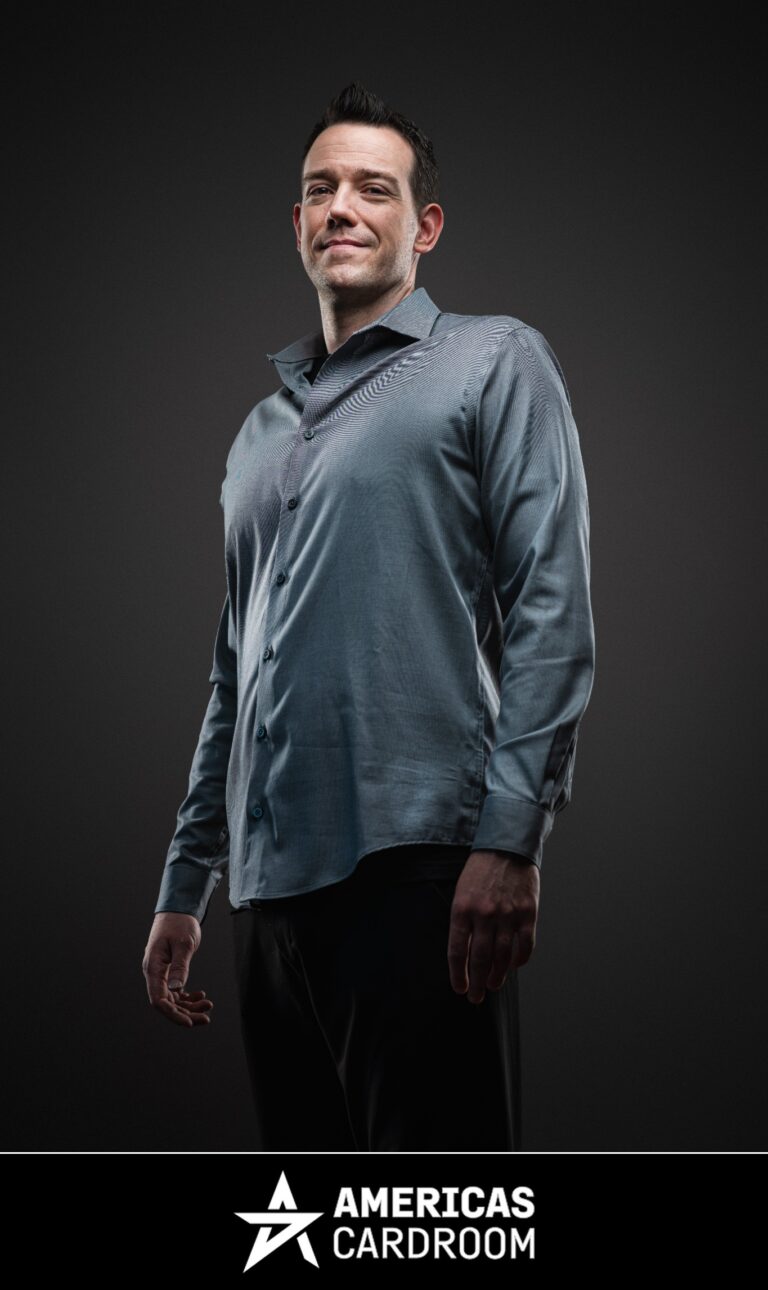 A medium shot of Jeff Boski, an ACR poker pro, smiling and looking to his left. He is wearing a long-sleeved grey button-up shirt and black pants, against a dark background.