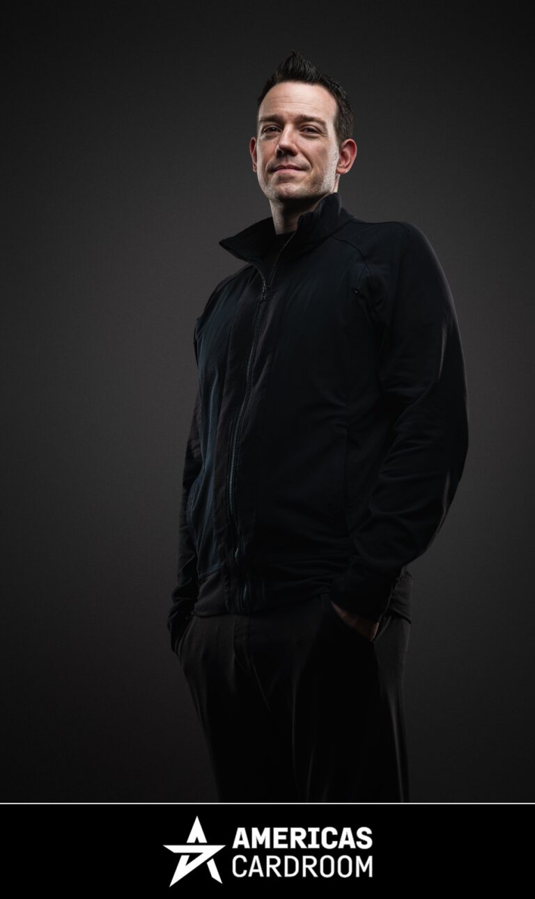 A medium shot of Jeff Boski, an ACR poker pro, looking up and to his left with a slight smile. He is wearing a black zip-up jacket and black pants, with his hands in his pockets, against a dark background.