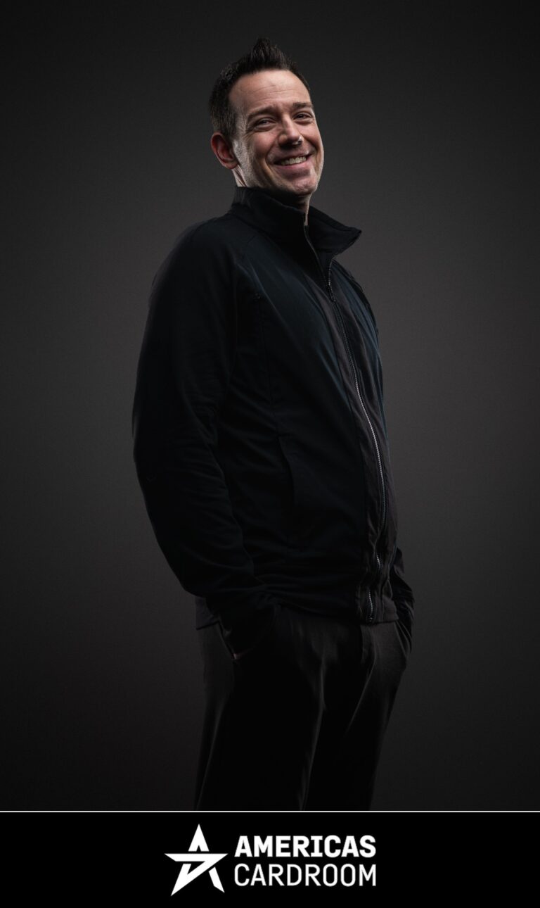 A medium shot of Jeff Boski, an ACR poker pro, smiling and looking to his left, wearing a black zip-up jacket and black pants, with his hands in his pockets, against a dark background.