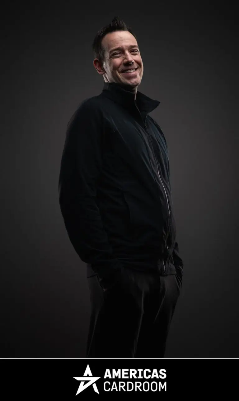 A medium shot of Jeff Boski, an ACR poker pro, smiling and looking to his left, wearing a black zip-up jacket and black pants, with his hands in his pockets, against a dark background.