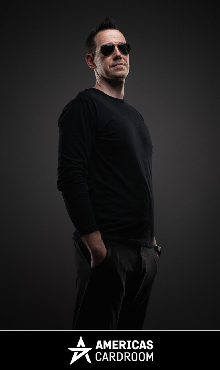A medium shot of Jeff Boski, an ACR poker pro, wearing sunglasses and a black long-sleeved shirt with black pants, looking intently to his right with his hands in his pockets, against a dark background.