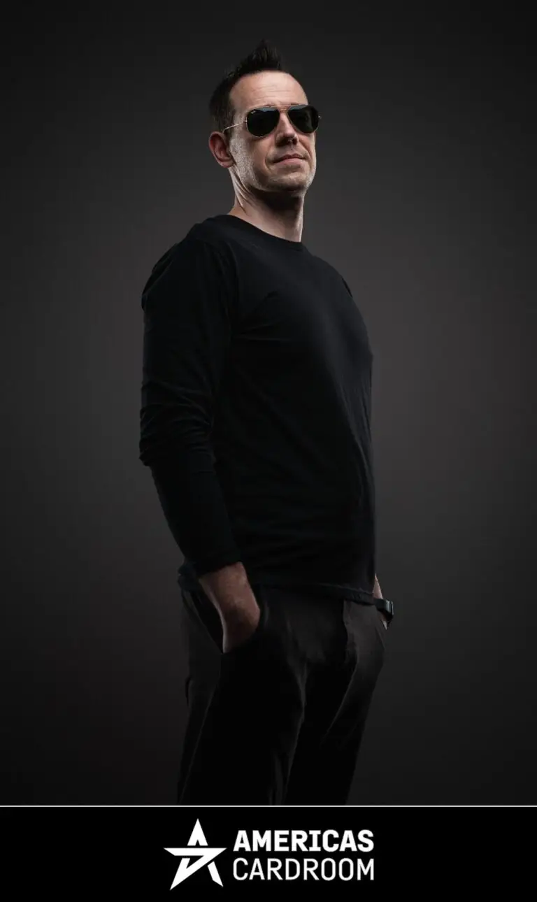 A medium shot of Jeff Boski, an ACR poker pro, wearing sunglasses and a black long-sleeved shirt with black pants, looking intently to his right with his hands in his pockets, against a dark background.