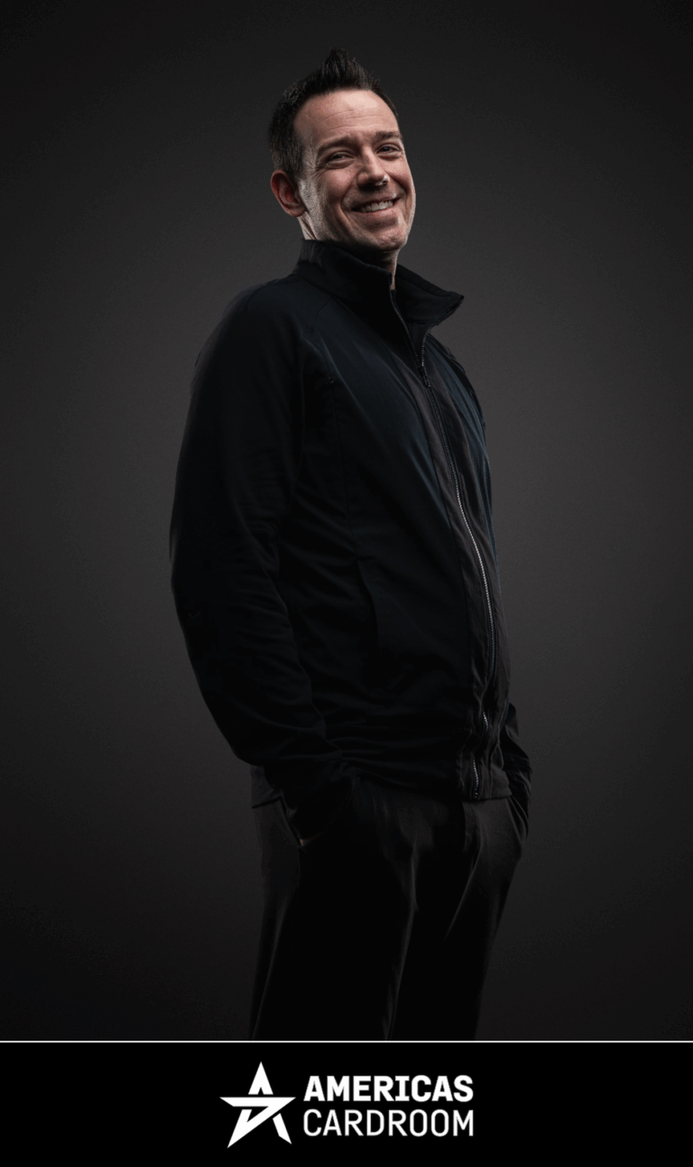 A medium shot of Jeff Boski, an ACR poker pro, smiling and looking to his left, wearing a black zip-up jacket and black pants, with his hands in his pockets, against a dark background.