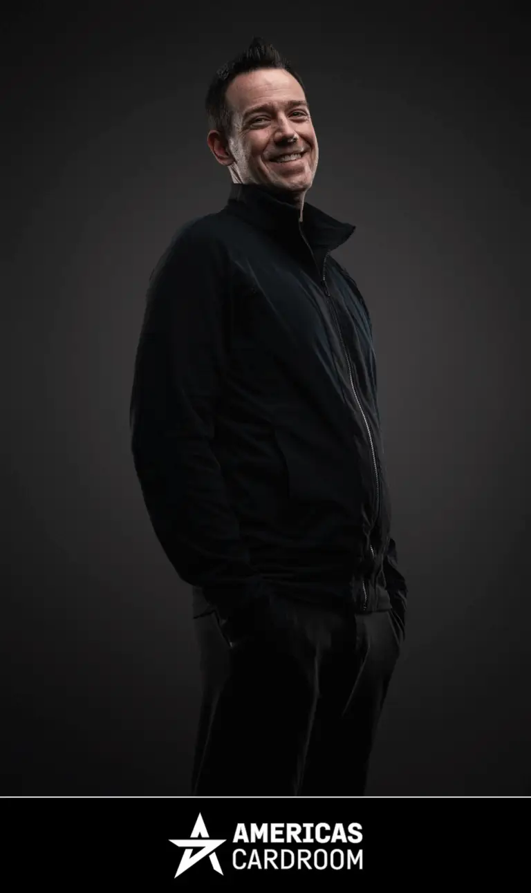 A medium shot of Jeff Boski, an ACR poker pro, smiling and looking to his left, wearing a black zip-up jacket and black pants, with his hands in his pockets, against a dark background.