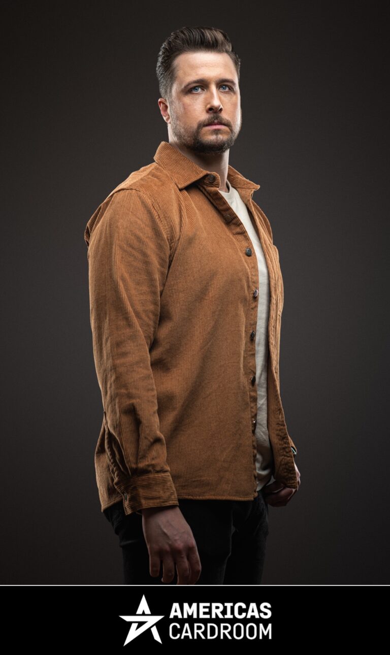 A medium shot of Jon Pardy, an ACR poker pro, with a beard, looking intently forward. He is wearing an open brown corduroy collared shirt over a light-colored t-shirt and dark pants, against a dark background.