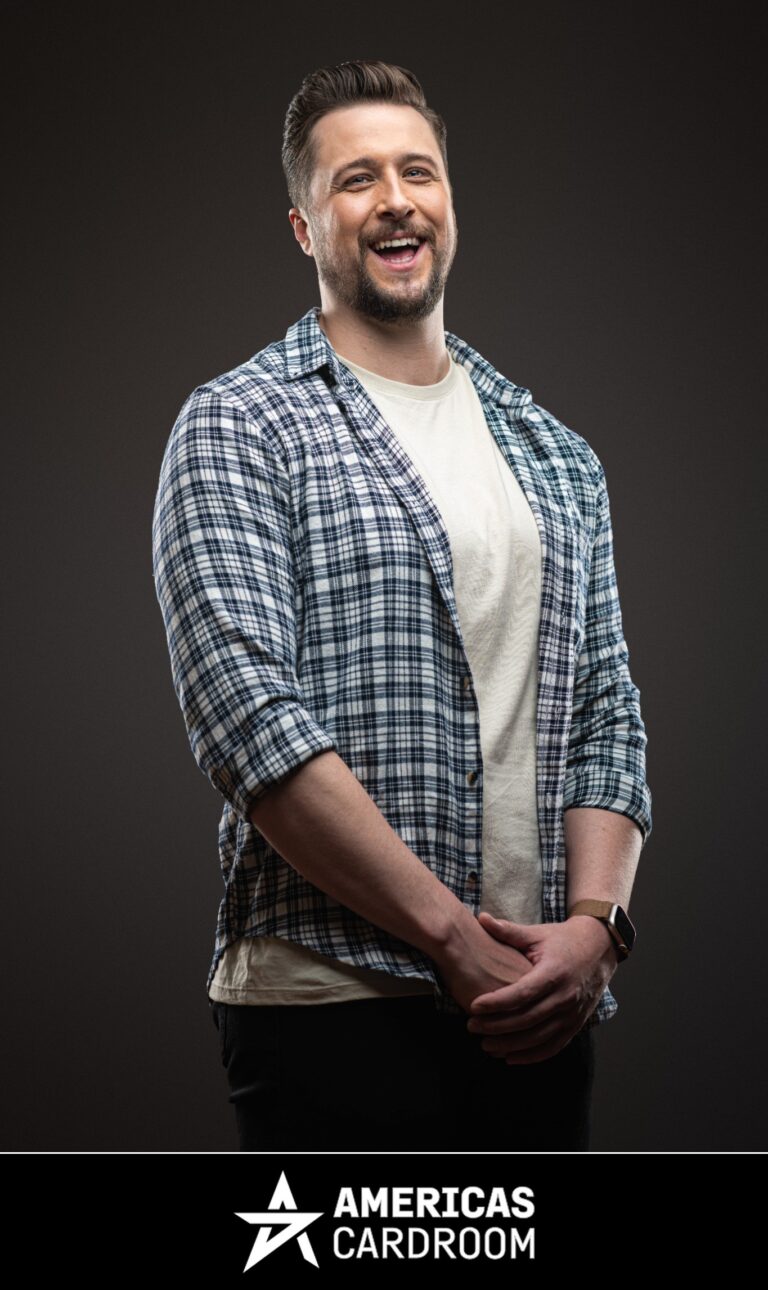 A medium shot of Jon Pardy, an ACR poker pro, with a beard, laughing with his mouth open and looking to his left. He is wearing a blue and white plaid button-up shirt open over a light-colored t-shirt, and dark pants, with his hands clasped in front of him, against a dark background.