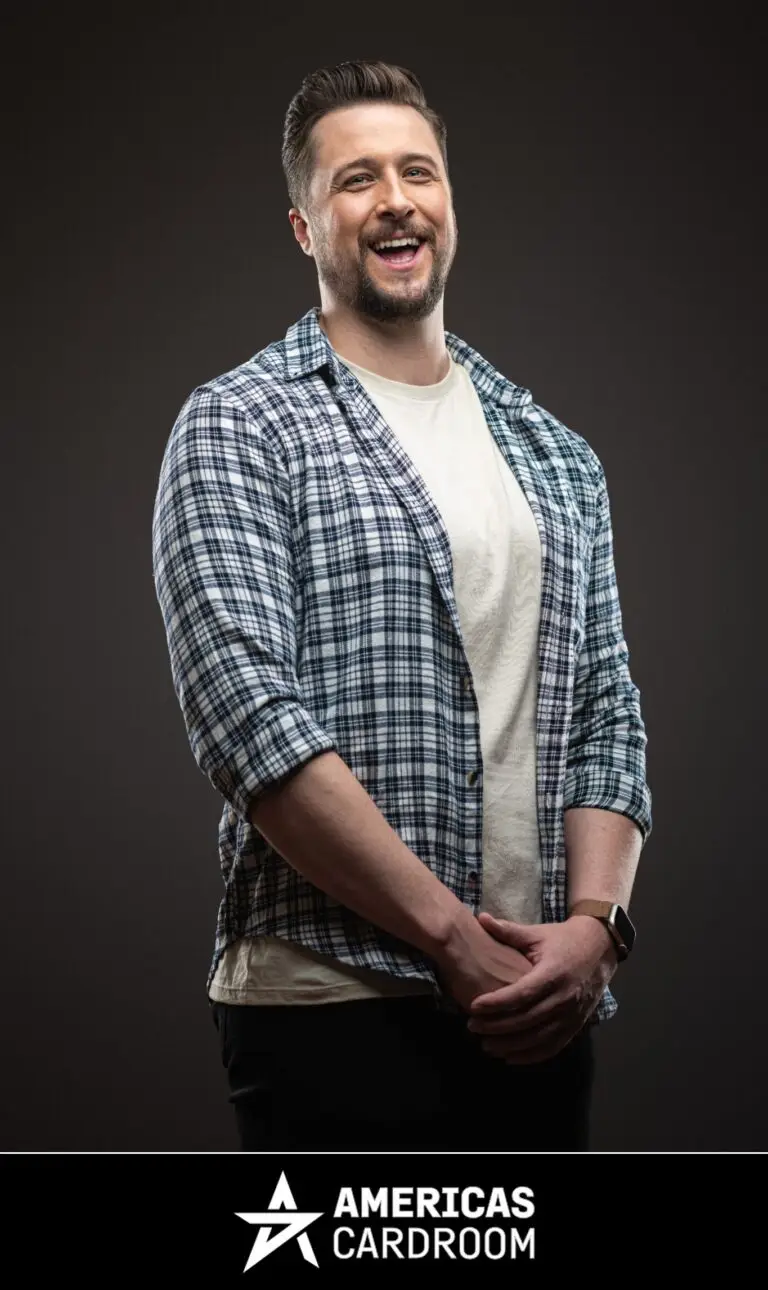 A medium shot of Jon Pardy, an ACR poker pro, with a beard, laughing with his mouth open and looking to his left. He is wearing a blue and white plaid button-up shirt open over a light-colored t-shirt, and dark pants, with his hands clasped in front of him, against a dark background.