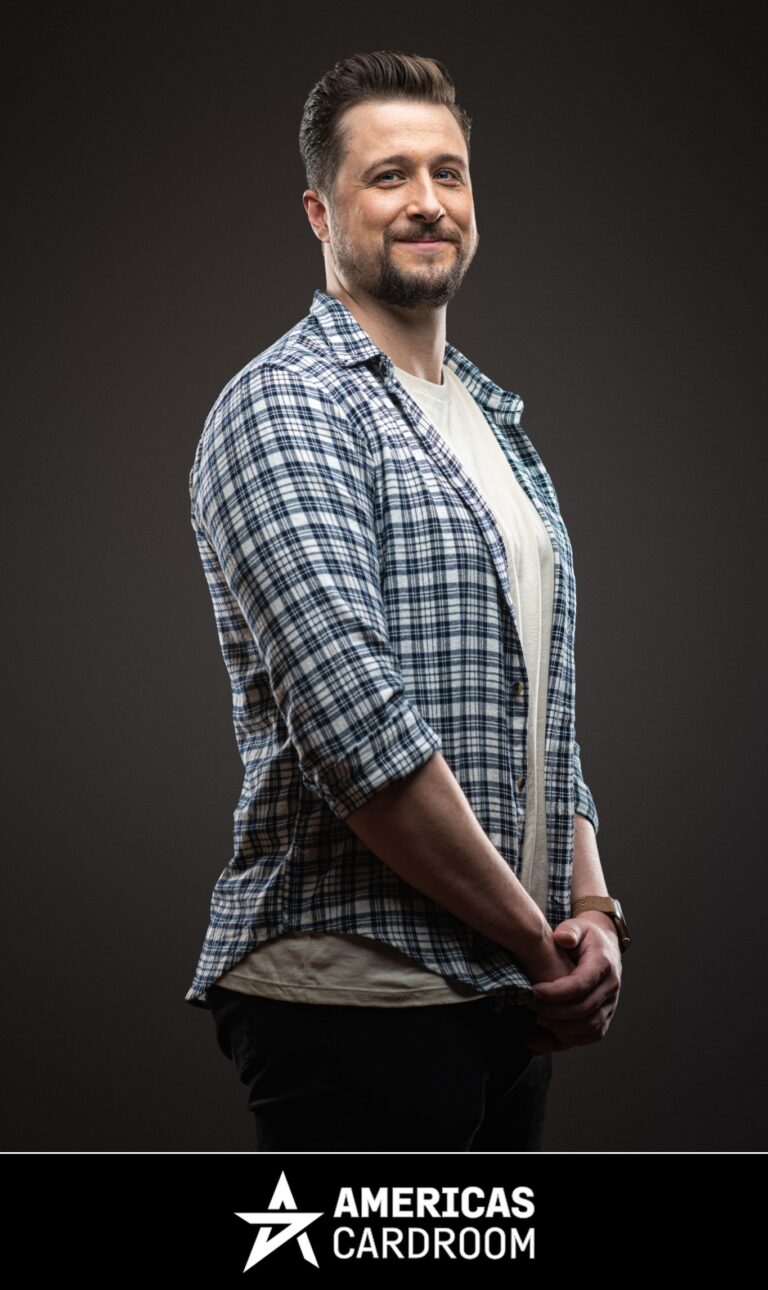 A medium shot of Jon Pardy, an ACR poker pro, with a beard, smiling and looking to his left. He is wearing a blue and white plaid button-up shirt open over a light-colored t-shirt, and dark pants, with his hands clasped in front of him, against a dark background.