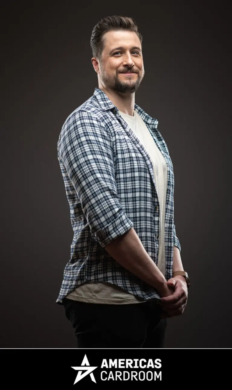 A medium shot of Jon Pardy, an ACR poker pro, with a beard, smiling and looking to his left. He is wearing a blue and white plaid button-up shirt open over a light-colored t-shirt, and dark pants, with his hands clasped in front of him, against a dark background.