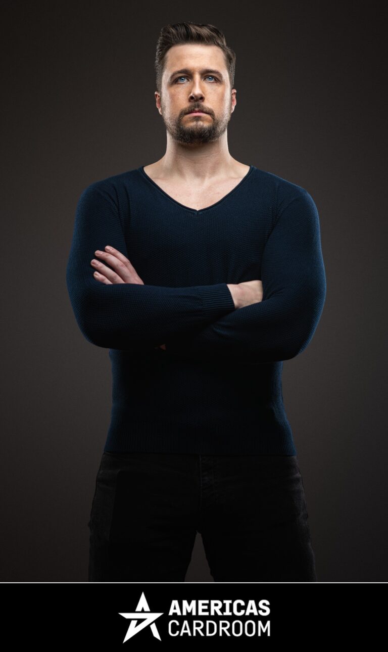 A medium shot of Jon Pardy, an ACR poker pro, with a beard, looking intently forward with his arms crossed. He is wearing a dark blue V-neck long-sleeved shirt and dark pants, against a dark background.