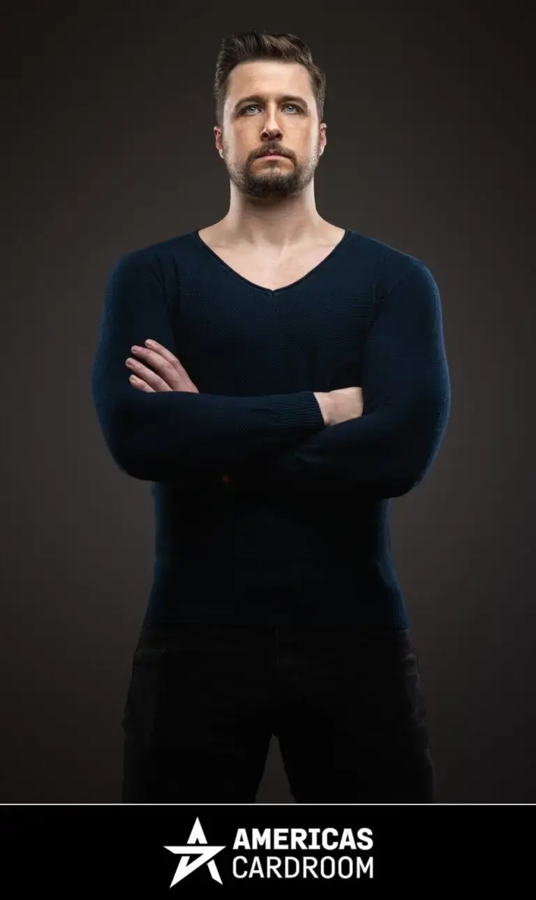 A medium shot of Jon Pardy, an ACR poker pro, with a beard, looking intently forward with his arms crossed. He is wearing a dark blue V-neck long-sleeved shirt and dark pants, against a dark background.