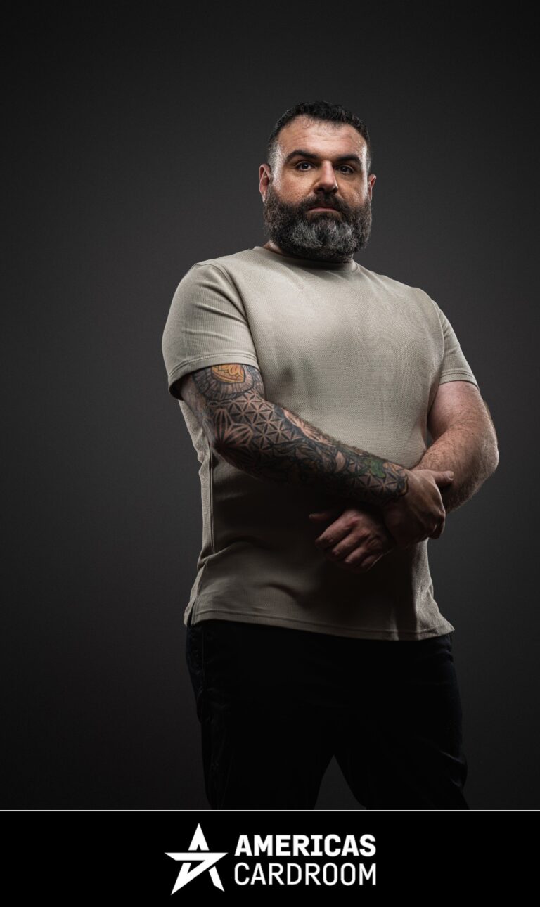 A medium shot of Jon Van Fleet, an ACR poker pro, with a full beard, looking directly at the camera with a serious expression. He is wearing a light grey t-shirt that reveals a full sleeve of tattoos on his left arm, and black pants, against a dark background.