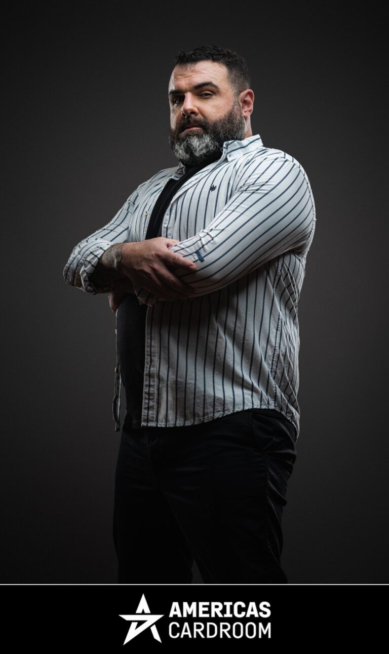A medium shot of Jon Van Fleet, an ACR poker pro, with a full beard, looking intently towards the camera with his arms crossed. He is wearing a light-colored button-up shirt with dark vertical stripes over a black t-shirt and black pants, against a dark background.